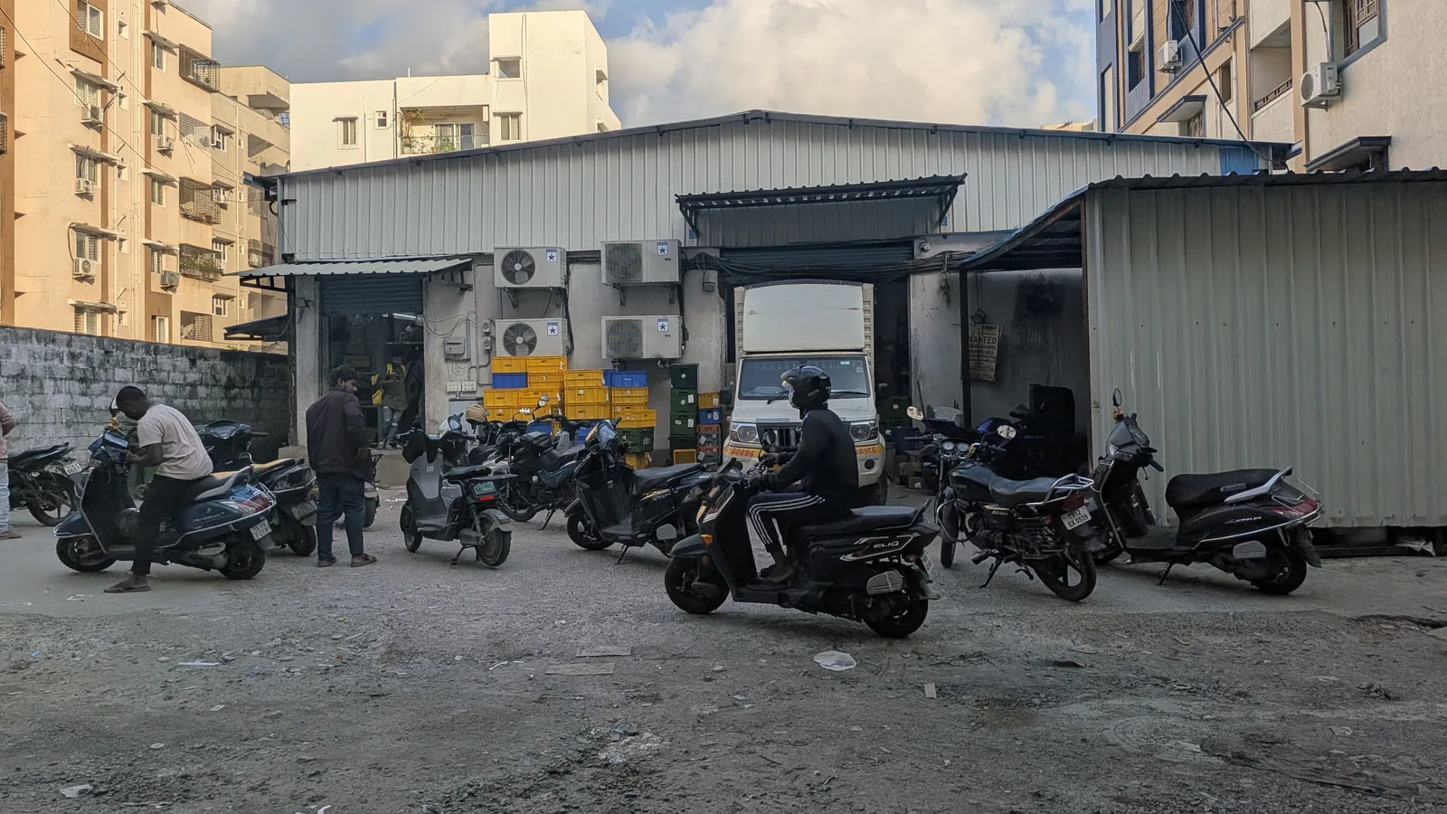 Delivery riders and a loading truck outside a small quick-commerce warehouse in Hyderabad, with stock crates stacked inside
