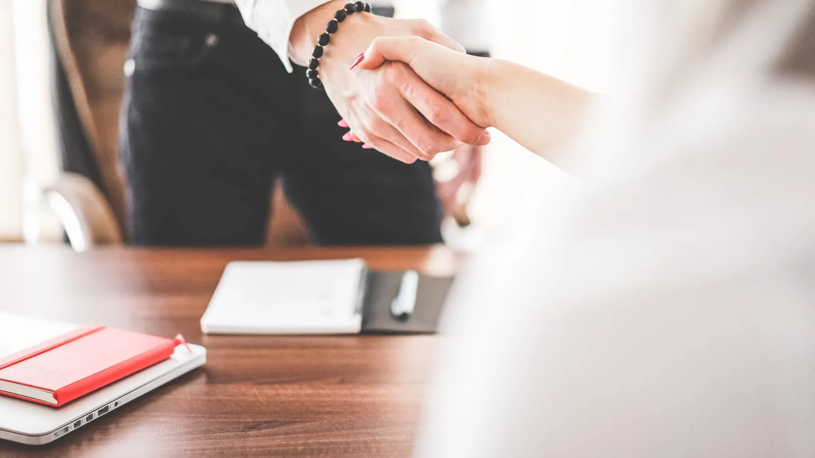 Two people shaking hands across an office desk with a laptop and notebook, closing a sales deal