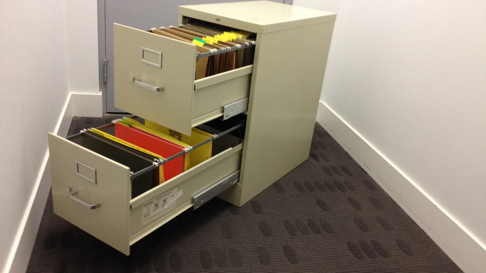 A beige metal filing cabinet with two open drawers full of hanging file folders in an office corridor