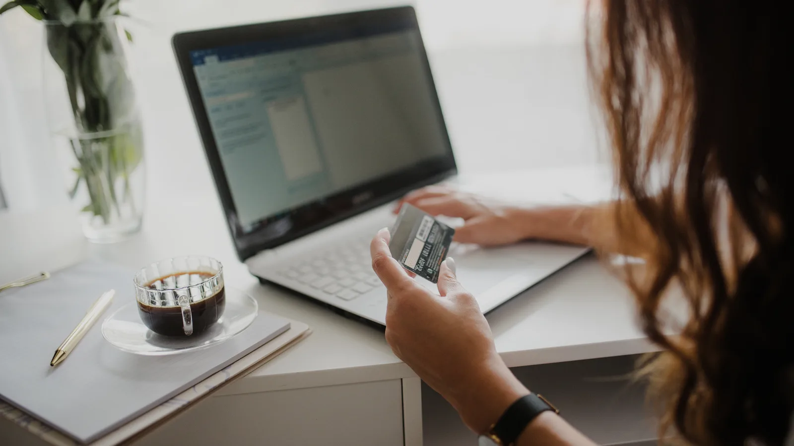 A person holding a payment card while using a laptop at a desk with a cup of coffee